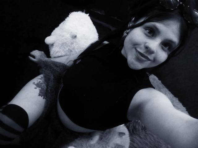 Sophie Hill, con piccoli seni, indossa una maglietta nera, ha un tatuaggio sul braccio destro, sorride alla fotocamera, dietro di lei c'è un peluche bianco, è una foto in bianco e nero, tiene in mano il telefono, ha gli occhiali sulla testa..