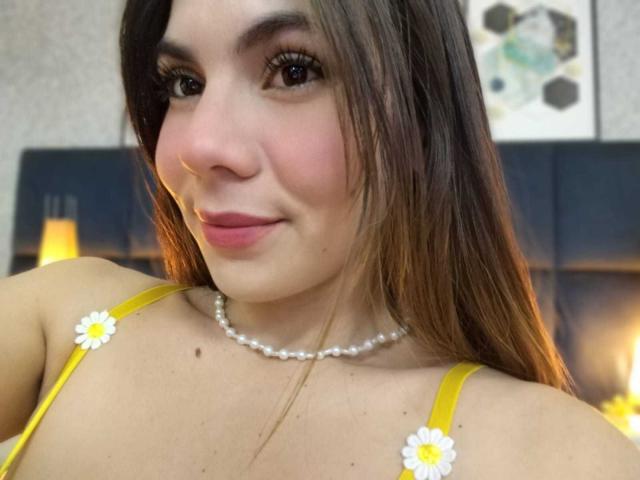 LindaPerez with brown hair, yellow dress, white daisy clips, pearl necklace, smiling, pink lips, soft makeup, visible tits, warm bedroom light, cute, attractive, medium shot.