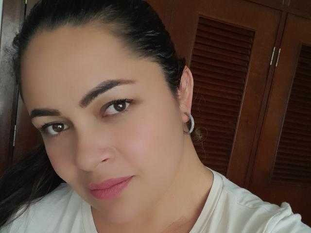 KristalNoa with medium brown skin, dark eyebrows, brown eyes, pink lipstick, white shirt, and silver hoop earring, smiling slightly, dark hair in a ponytail.