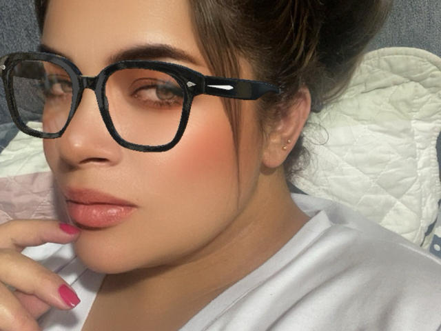 KristalNoa has glasses on, pink lipstick, and pink nail polish, with her hand near her mouth, showing off her cute, slightly blushed face and soft brown hair.