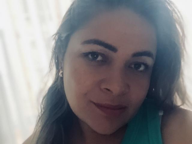 KristalNoa has dark brown eyebrows, green eyes, and a subtle smile. She's wearing a green top and has wavy brown hair, with a small nose ring on her left side.