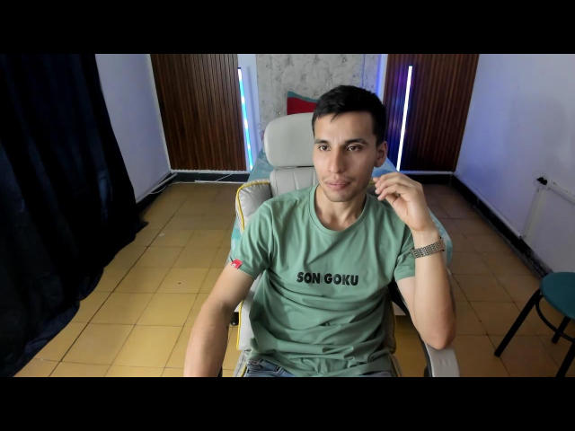 SexyCaicer with short black hair, wearing a green "SON GOOKU" t-shirt, sitting in a chair, holding earbuds, looking at camera, tile floor, wooden wall, and chair visible.