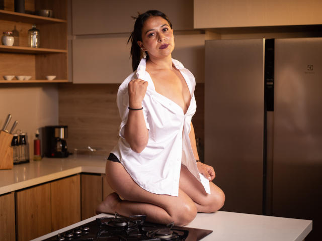 AfroditaLaDiosa kneels on a kitchen counter, wearing an unbuttoned white shirt revealing black panties. She has a medium build, dark hair in a ponytail, and is holding the shirt's collar with a slight smile. The background includes wooden shelves, a knife set, and a stainless steel fridge. The lighting is warm, highlighting her skin tone. The image has a watermark in the bottom left corner.