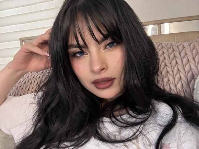 Close-up of ClariceMontclair with long, straight black hair and bangs, blue eyes, and dark red lipstick. She has light skin with a few small freckles on her cheeks. ClariceMontclair is wearing a white shirt and is resting her right hand in her hair. The background has a beige, textured pillow and white horizontal blinds. Her makeup is bold, with thick eyeliner and long lashes. The image is sharp, with soft lighting.