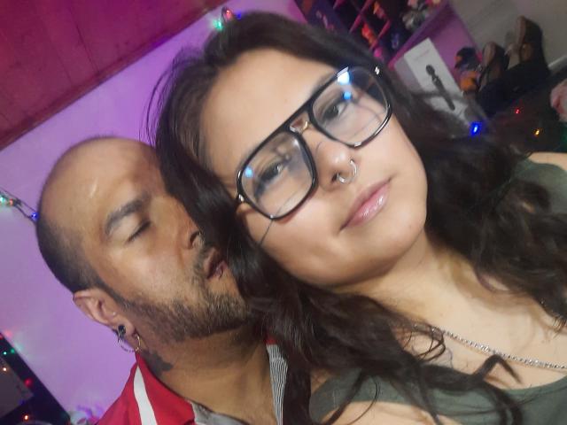 SektethLilith with glasses and nose ring, brown hair, and green top, leaning on bald, bearded man with red jacket. He's nuzzling her neck, with colorful lights.