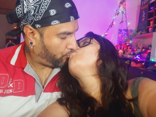SektethLilith, a man with a black bandana and red-and-white shirt, kisses SektethLilith, a woman with dark hair and glasses, on the lips. He's got a nose ring, she's got tits.