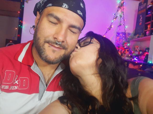 SektethLilith with glasses, kissing the nose of SektethLilith in a red and white shirt, black bandana, nose ring, and beard, in a cozy, colorful room.