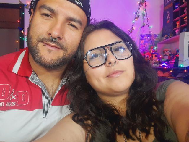 SektethLilith has a nose ring and is wearing glasses, standing next to a bearded man with a nose ring, both with dark hair, in a cozy indoor setting.