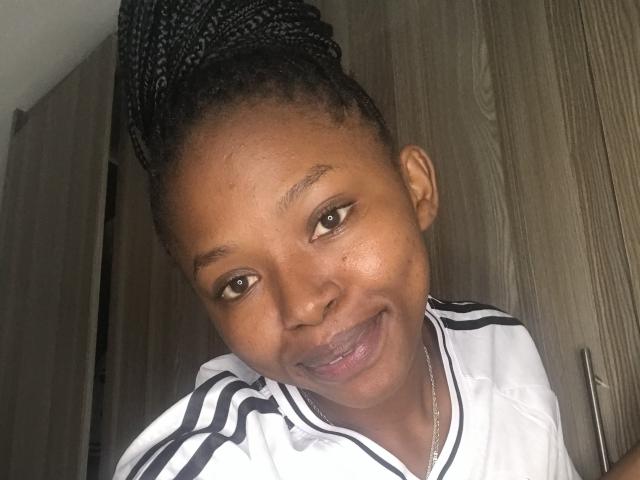ViolettaCleame has dark brown skin, braided black hair in a high bun, and is smiling with her tongue slightly out. She's wearing a white Adidas shirt.