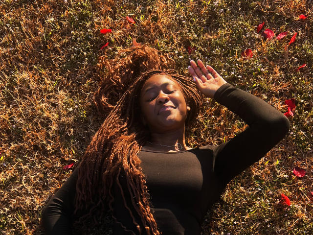 GiselleCumsForU, lying in grass with red petals, eyes closed, black long-sleeve shirt, arm raised, brown braided hair, sunlit, relaxed, small smile, soft light, natural setting.