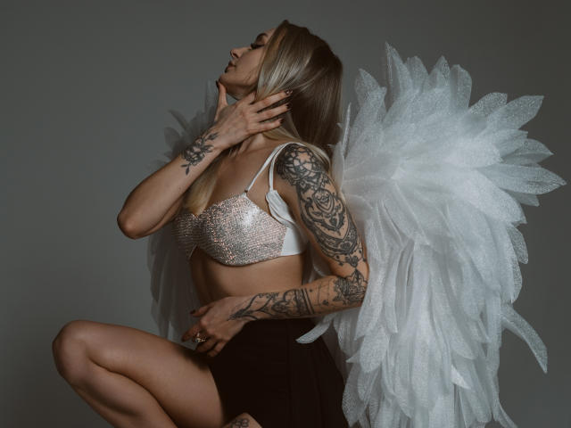 CandyGraces kneels with white, fluffy angel wings. She's wearing a sparkling white bra and a black skirt. Her blonde hair is down, and she's touching her neck. Her left arm, covered in detailed black tattoos, rests on her knee. The photo has a dark, moody tone with sharp contrasts. The wings are textured and fluffy, adding a soft, ethereal element to the image.