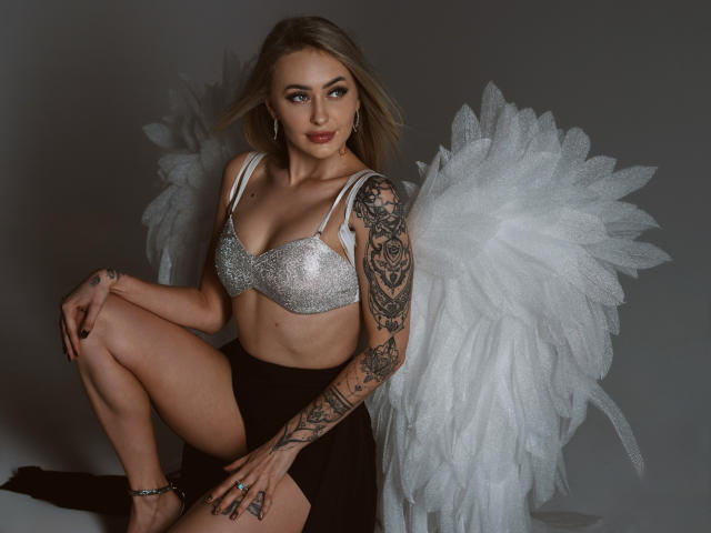 CandyGraces with silver sparkly tits, white feather wings, tattooed arm, and black skirt, squatting, looking at camera, blonde hair, gray background, earrings, ankle bracelet.