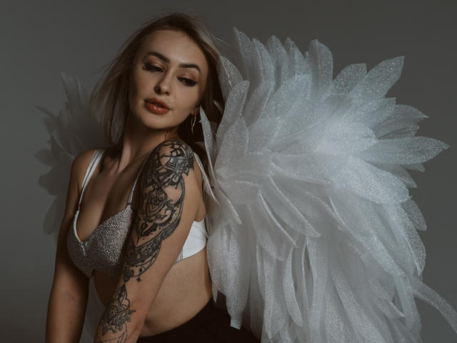 CandyGraces, a blonde woman with fair skin, wears a white bra and large, glittery, white feathered wings. She has a detailed black tattoo on her right arm and is looking down with a slightly open mouth. Her makeup is bold with dark eyeliner and glossy lips. The image is lit with a soft shadow, highlighting her wing and arm tattoo. Slightly angled, she stands against a gray background.