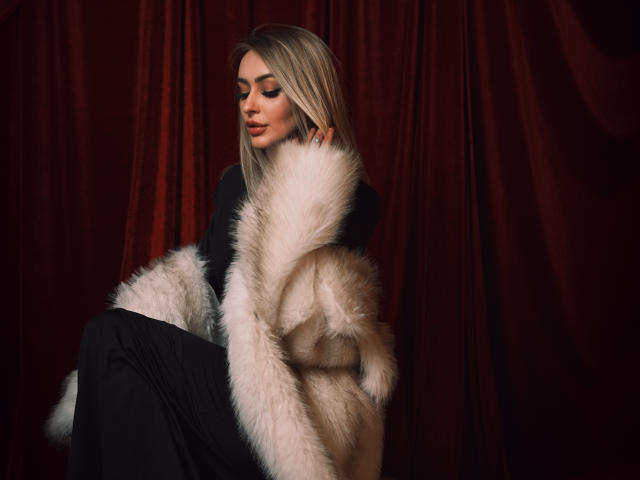 CandyGraces with blonde hair, black top, and fluffy white fur coat, sitting with eyes closed, pouty lips, and black pants, looking sexy and stylish.