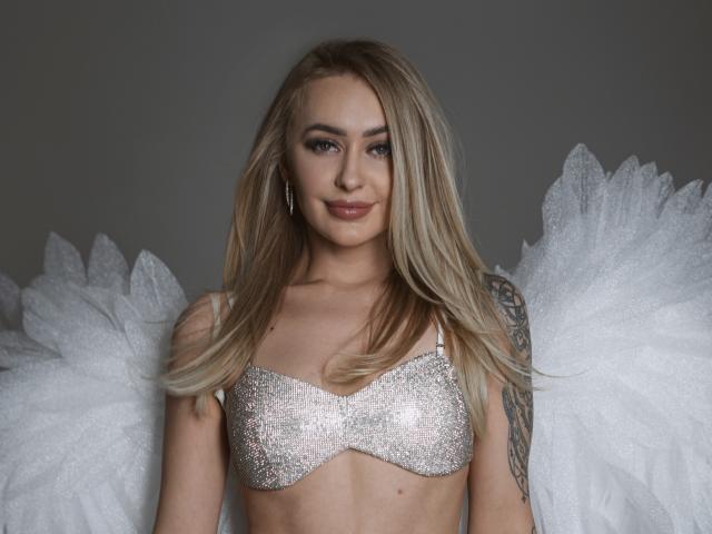 CandyGraces, a blonde woman with light skin and blue eyes, wears a sparkly silver bra and white, textured angel wings. She has long hair, subtle makeup, and a tattoo on her right arm. Her confident expression is enhanced by small, silver hoop earrings. The image uses soft lighting, emphasizing her smooth skin and the glittery texture of her bra. The wings are fluffy and white, contrasting with her sleek hair. The overall style is elegant and ethereal.