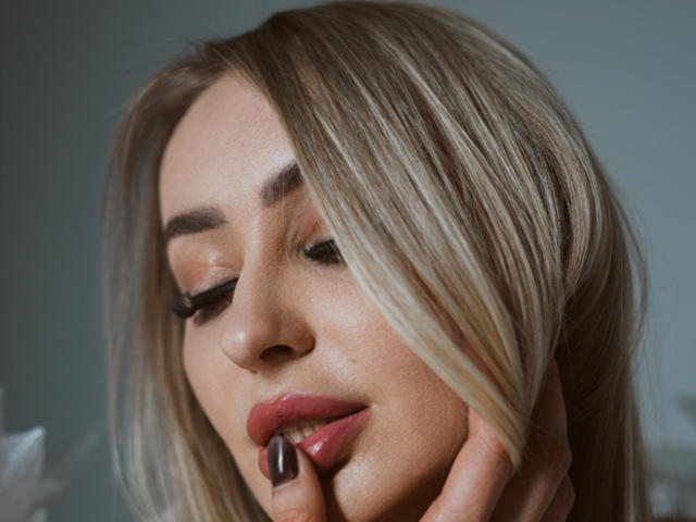 Close-up of CandyGraces with blonde hair, fair skin, and subtle makeup. Her right hand, with black-painted nails, gently touches her lower lip. She has soft pink lips and dark eyeliner. Her eyes are closed, and her head is slightly tilted. The lighting is soft, highlighting her smooth skin and hair's natural shine. The image has a calm, intimate feel with a slight shadow on her left cheek. The photograph focuses on her face and hand, with a shallow depth of field.