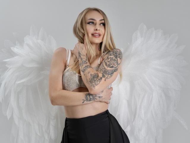CandyGraces with long blonde hair, wearing a sparkly bra, black skirt, and white angel wings, has tattoos on her arms and a cute smile. Boobs are visible.
