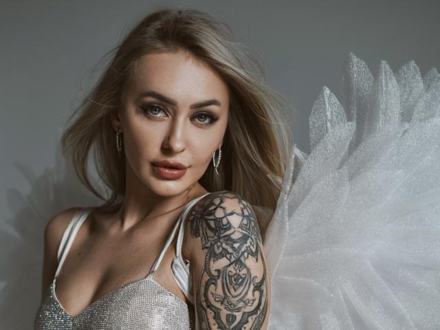 CandyGraces has long blonde hair, light skin, and intense blue eyes. She wears a sparkly white dress with thin straps and glittery earrings. Her right shoulder, adorned with a detailed black tattoo of roses and geometric patterns, is prominent. White, glittery wings extend from her back, adding a celestial vibe. Her makeup is subtle but enhances her natural beauty. The image is sharp and focused on her upper body.