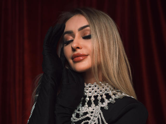 CandyGraces with long blonde hair, black gloves, and a jeweled black top, eyes closed, smiling, touching her head. Red velvet background. Boobs partially visible under top.