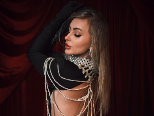 CandyGraces with long brown hair, black gloves, and pearls draped over her bare back, showing her tits. She's got a serious expression and dark red velvet curtains.