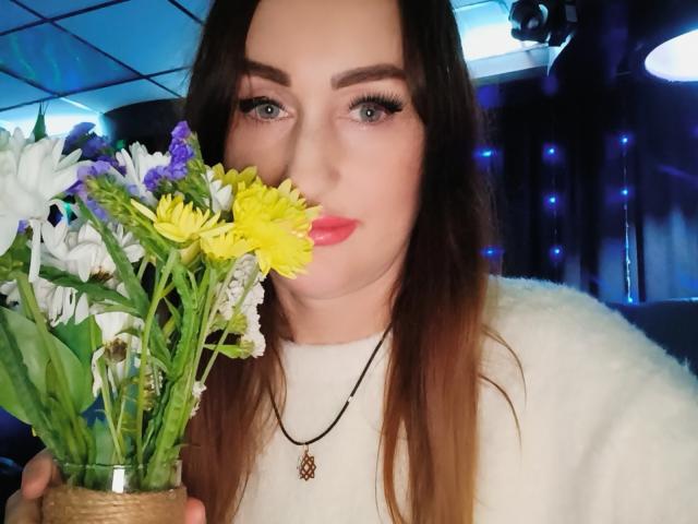 FreyaHott69 mit langen braunen Haaren, blauen Augen und pinkem Lippenstift, hält einen Blumenstrauß aus gelben, weißen und lila Blumen, trägt einen weißen, flauschigen Pullover und eine Kette mit einem Pentagramm..