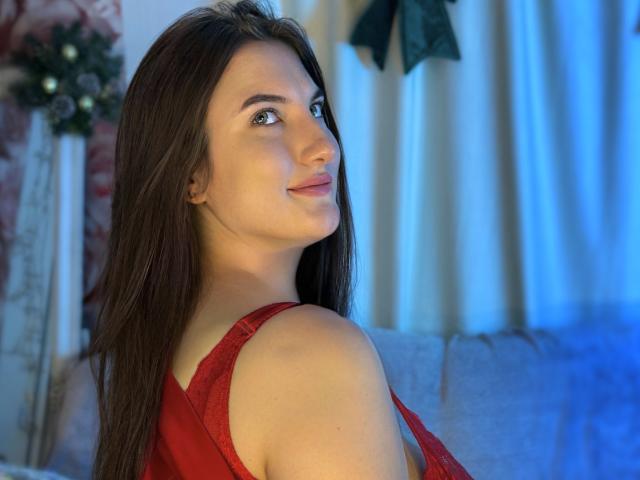 StephieRouse with long brown hair, red sleeveless top, looking over shoulder, smiling, blue light background, visible ass, light skin, soft makeup, cute, relaxed.