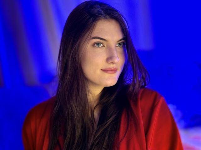 StephieRouse has long brown hair, blue eyes, and a slight smile. She wears a red shirt, and the blue background adds a cool contrast to her warm skin.