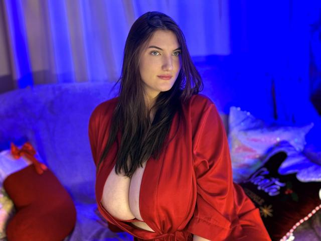 StephieRouse with huge tits in a red silky robe, long brown hair, blue lighting, sitting on a couch with red and white Christmas sacks.