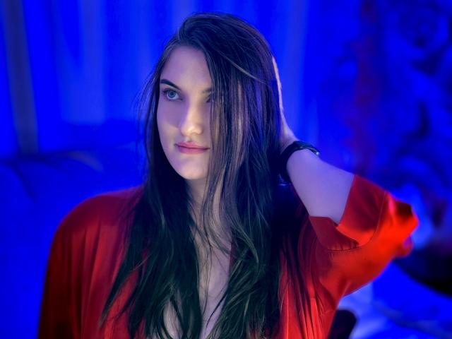 StephieRouse with long black hair, blue eyes, red shirt, hand in hair, slight smile, blue lighting, wearing black wristband, subtle makeup, sexy, confident, photograph, upper body.