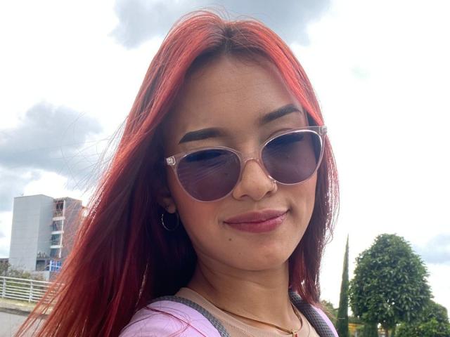 SashaHans, con cabello rojo, usando gafas de sol, una camisa morada y aros grandes, sonriendo ligeramente. Tiene una cara linda y senos pequeños. El cielo está nublado..