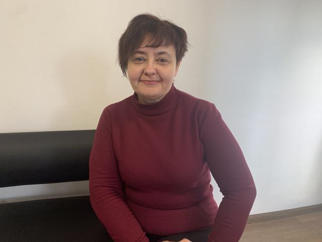 JustinaMavka, short brown hair, smiling, wearing a red turtleneck sweater, sitting on a black bench, hands on her lap, wooden floor, white wall in background.