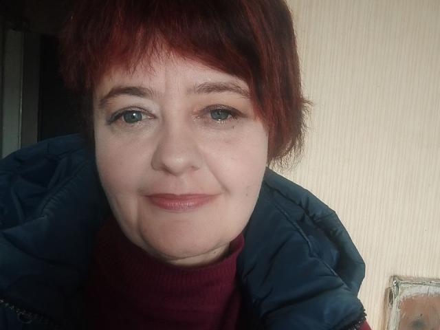 JustinaMavka with red hair, green eyes, and a slight smile, wearing a dark blue jacket and red turtleneck, showing a bit of her ass in the background.