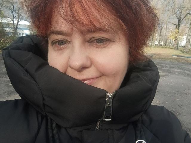 JustinaMavka's face, showing red, messy hair, black puffy jacket with zipper, and a slight smile, with her nose and lips slightly visible, standing outside in a park.