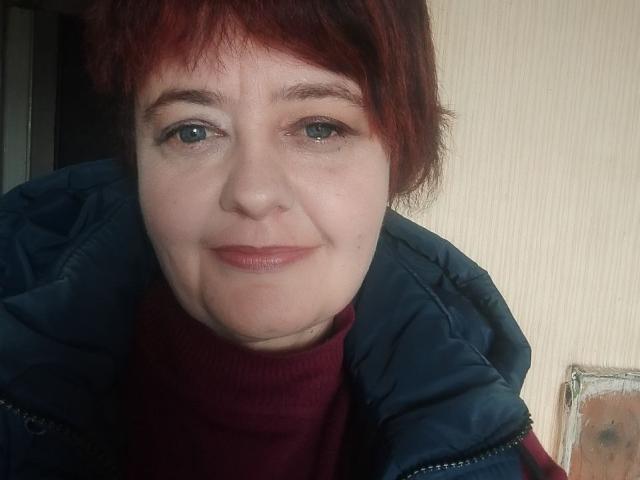 JustinaMavka with short red hair, green eyes, and light skin, wearing a navy blue jacket and red turtleneck, smiling slightly. She has a small nose and subtle makeup.