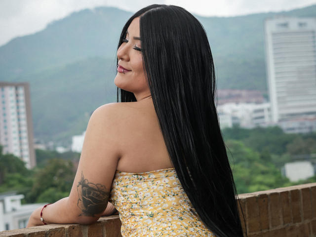 ValkiMoore with long black hair, yellow floral dress, bare shoulders, tattoo on right arm, smiling over brick ledge, city and green hills in background. Boobs not visible.