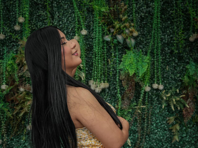 ValkiMoore with long black hair, eyes closed, smiling, holding her tits, wearing a yellow patterned dress, standing against a green wall with hanging plants.