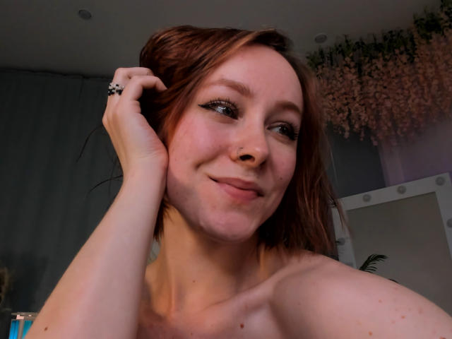 LilianKroft has light skin, red hair, and is smiling with a slight nose piercing, touching her hair with a black ringed finger, showing bare shoulders and subtle freckles.