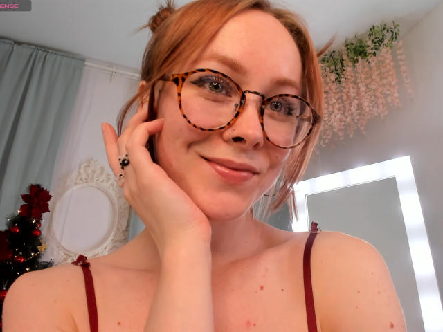 LilianKroft has red hair in buns, tortoiseshell glasses, and a cute smile, touching her cheek, wearing a red strap dress, showing her tits, in a cozy room.