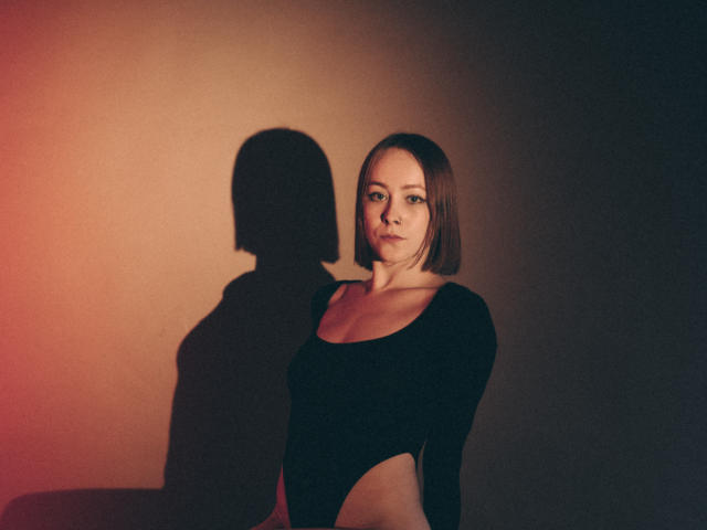 LilianKroft with shoulder-length brown hair, wearing a black long-sleeve bodysuit, showing her bare ass, shadow on the wall, soft orange-red light on the left.