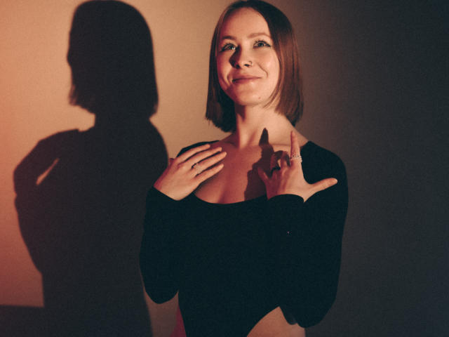 LilianKroft with short brown hair, wearing a black long-sleeve top, hands near chest, shadow on wall, smiling softly, light brown skin, subtle makeup, confident pose.