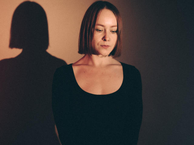 LilianKroft with a bob haircut, nose ring, and black top, standing with her shadow on the wall, looking down, showcasing her small tits and gentle facial features.