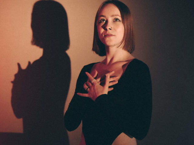 LilianKroft with short brown hair, black top, hands on chest, shadow of her with raised finger on left. Warm, dim lighting. Boob shadow, ring on finger, serious expression.