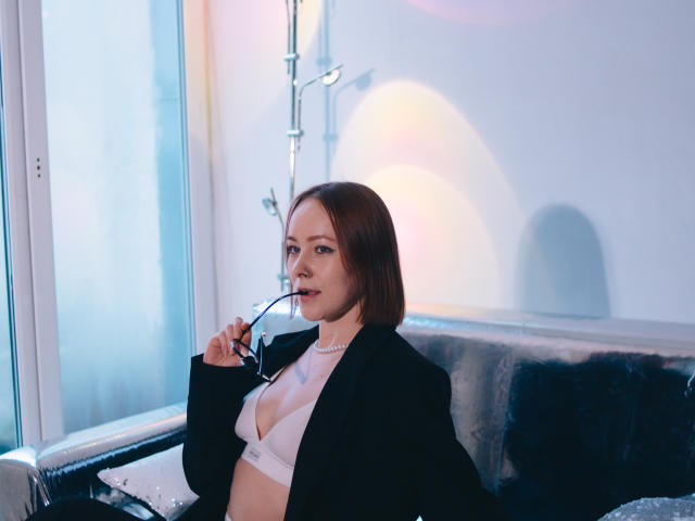 LilianKroft with short brown hair, wearing a black blazer and white bra, holding glasses, looking at camera, in a bright room with blue and pink light reflections.