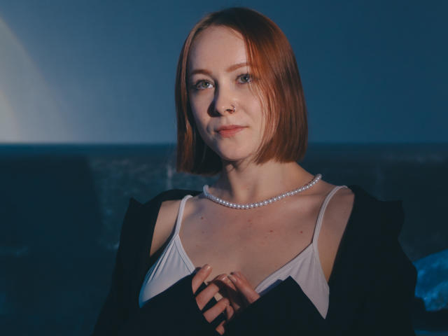LilianKroft with short red hair, pale skin, and a nose ring, wearing a white top and pearl necklace, hands together, dark blue light, slight smile, tits visible.