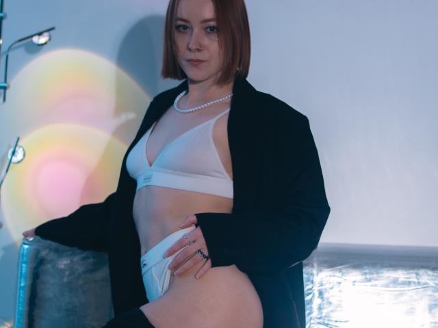 LilianKroft with short brown hair, white bra, and pearl necklace, wearing a black jacket, exposing her tits and white panties, standing in a blue-lit room, showing a confident look.
