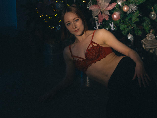LilianKroft with red lace bra, showing her tits, and black pants, leaning against a Christmas tree with pink ornaments, in dim lighting, looking at the camera.