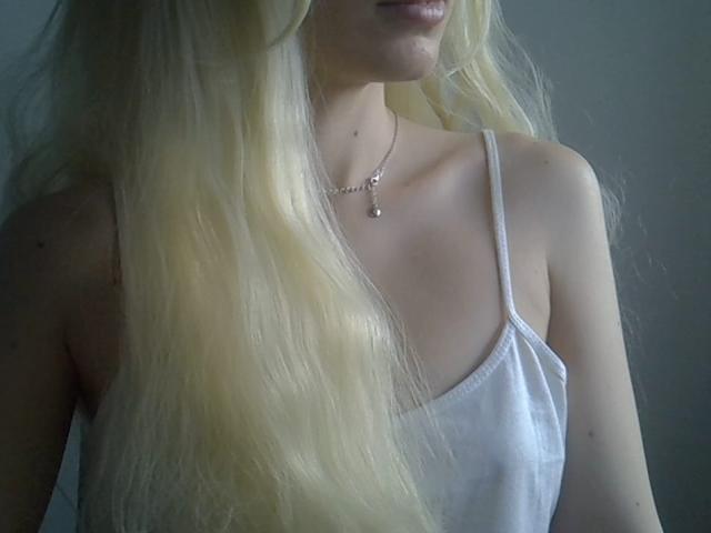 Close-up of DitaVira with long, wavy blonde hair covering her right shoulder. She wears a white spaghetti strap top and a delicate necklace with a small pendant. Her skin is light, and the lighting is soft, creating a gentle shadow on her left shoulder. The focus is on her hair and upper chest, with a subtle, natural texture to the hair. Slight lip shadow visible. The composition is simple and centered.