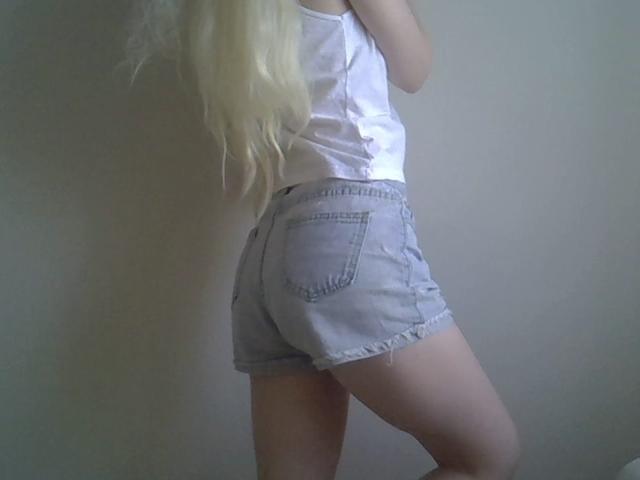 DitaVira with long blonde hair, wearing a white tank top and light blue denim shorts, standing sideways, showing her ass and upper thighs.
