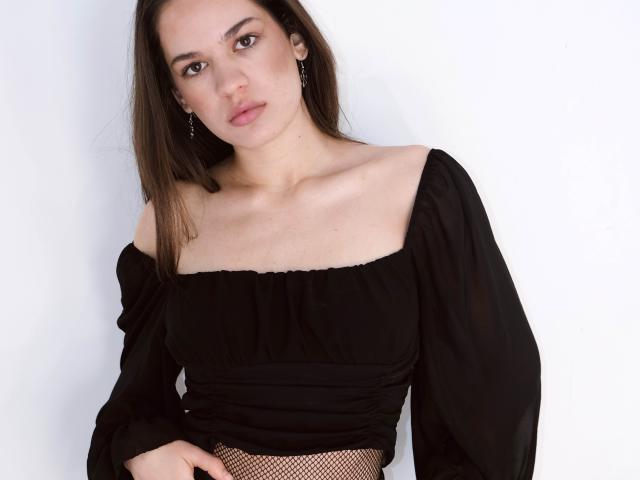 BeverlyHeart has brown hair, a pale skin tone, and is wearing a black off-shoulder top with fishnet underneath, showing a little of her tits. She's looking at the camera.