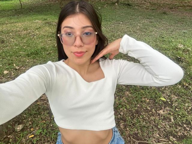 IsabelaPretty with glasses, white long-sleeve crop top, blue jeans, and pink lipstick, posing outdoors, hand near chin, showing her tits and flat stomach.